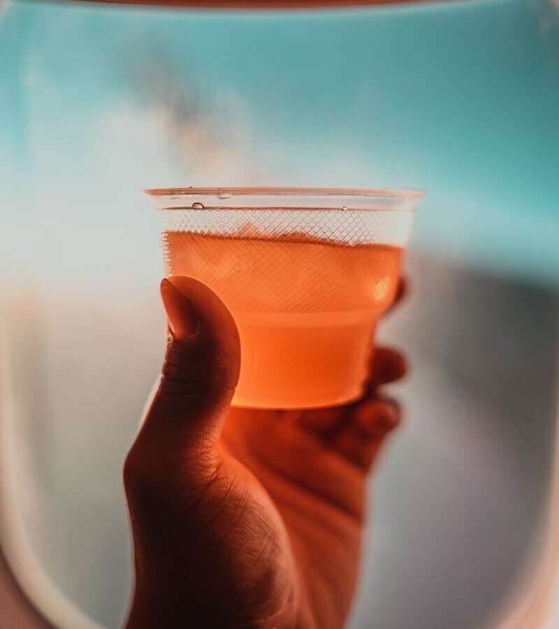 A close-up of a drink held by hand against an airplane window with a warm sky view.