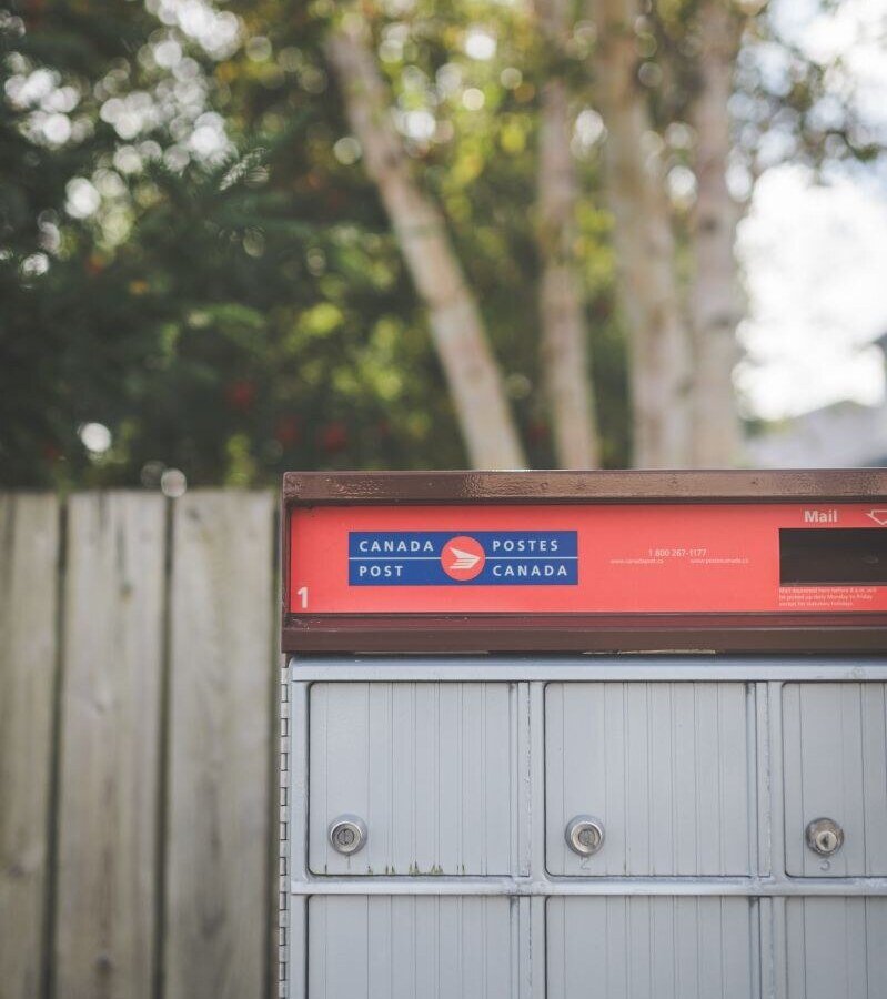 A Canada Post mailbox with colorful label outdoors with trees in the background.