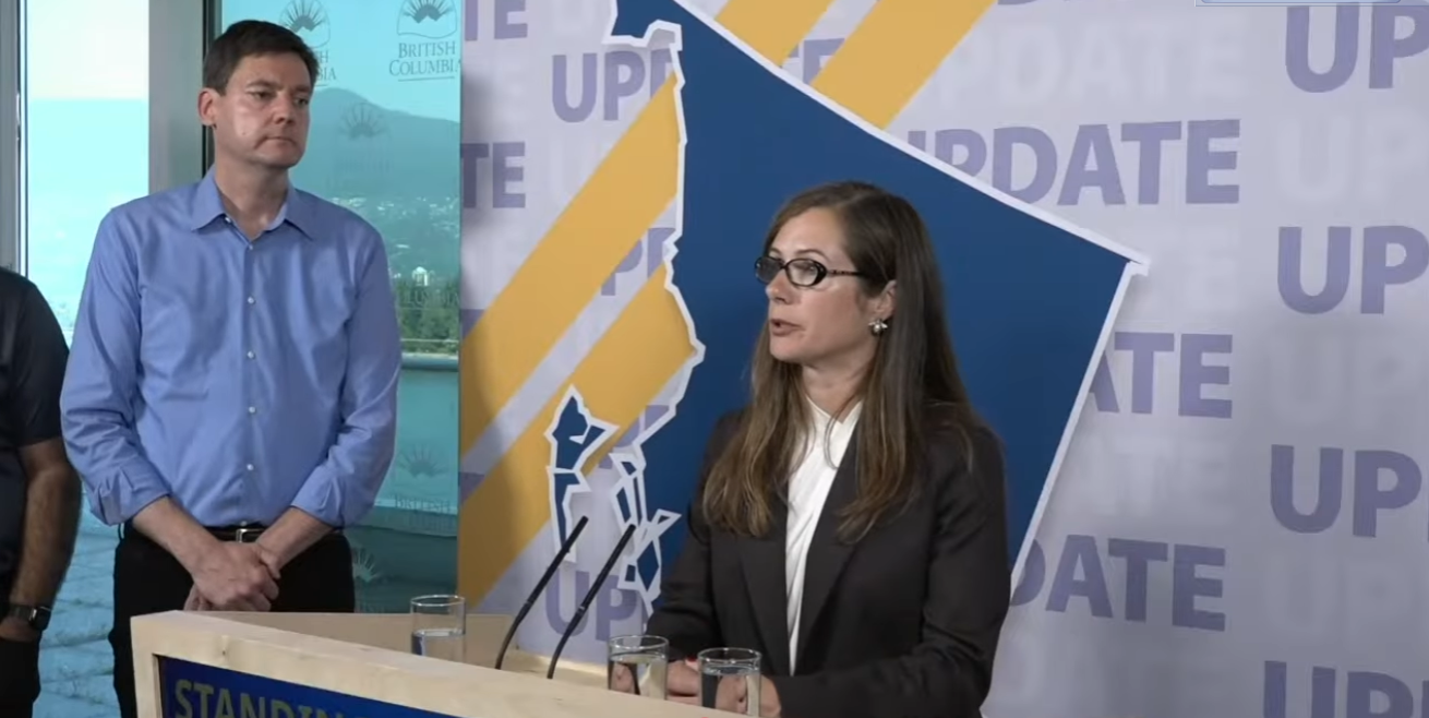 Premier David Eby stands at a wildfire update podium with two provincial ministers in frame during a British Columbia emergency briefing, with BC government “UPDATE” signage and a province map backdrop behind them.