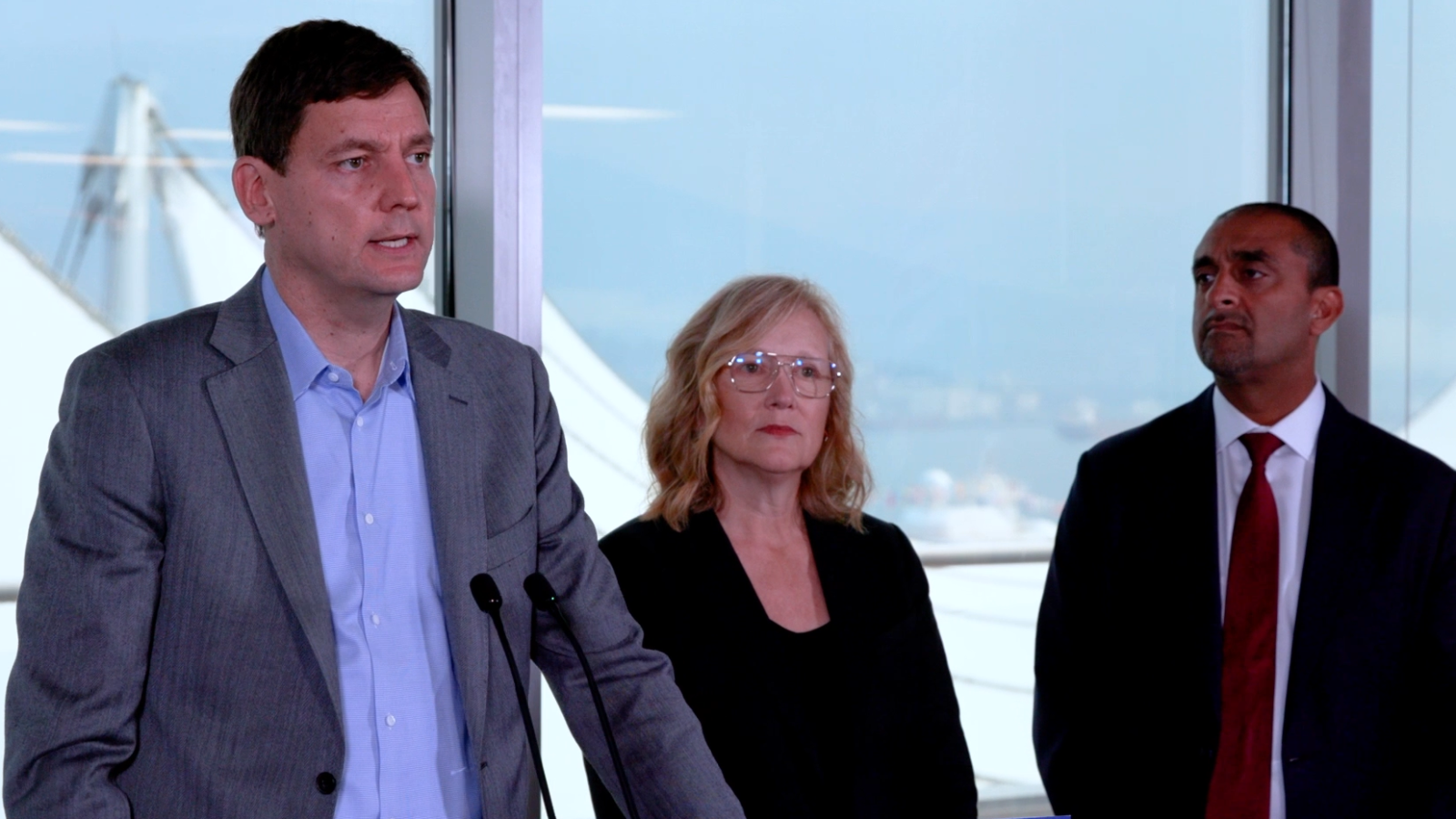 Premier David Eby speaking at a podium with Brenda Bailey and Ravi Kahlon standing behind him during a press conference