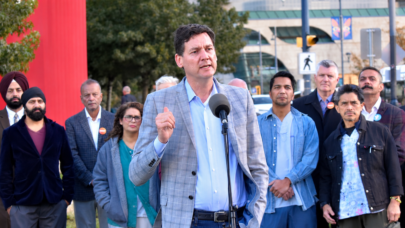 David Eby speaks into a microphone outdoors in Surrey, wearing a grey checked blazer and light blue shirt, with a group of people standing behind him.